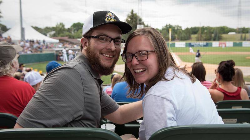 A man and woman smile at the camera with the baseball field in the background.