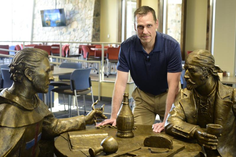 ​​​​​​​Sean Thorenson, the master illustrator of the new Marauders logo, stands next to the Monk and the Marauder sculpture in the Crow’s Nest Restaurant—the inspiration for his logo creation.