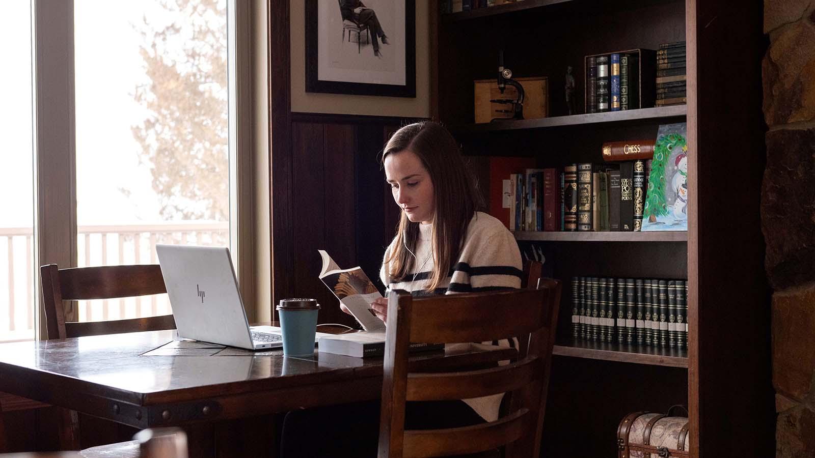 English student reading by window with coffee and laptop on table