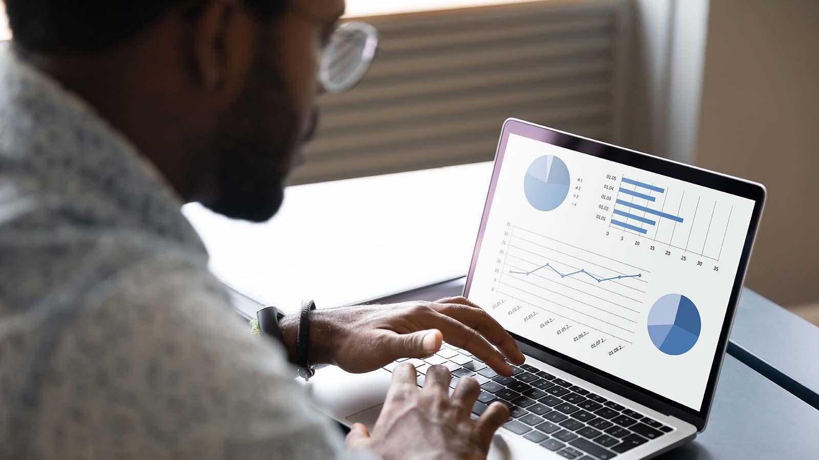 Focused young african american male professional working on project report by analyzing graphs and charts, sitting at table in office or home.